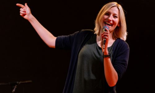 Sängerin Sarah Kaiser - Beitragsbild - Sarah Singt. Auf dem Bild sieht man Sarah Kaiser. Eine Sängerin mit blonden Haaren. Sie hat ein Mikrofon in der Hand und lächelt singend in die Kamera, so als hätte sie spaß.