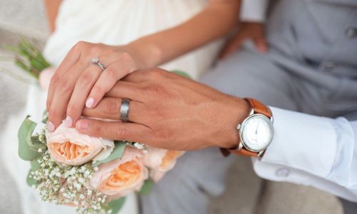 Hochzeitssängerin. Auf dem Bild sieht man die Hände eines Brautpaars, die auf einem Brautstrauß übereinander gelegt sind. An ihren Fingern, sieht man Ringe. Wahrscheinlich sind das die Eheringe.