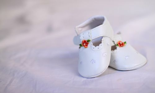 Sängerin für die Taufe - Cover-Bild. Auf dem Bild sieht man zwei weiße Baby Schuhe auf einer weißen Decke. Die Schuhe haben eine Blumenverzierung oben an der Öffnung der Schuhe. Die Blumenverzierung ist grün mit orangenen Blüten.